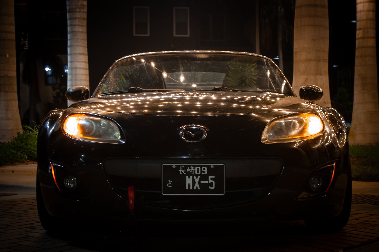 Mazda MX-5 Miata at night with headlights on under string lights — cinematic JDM portrait